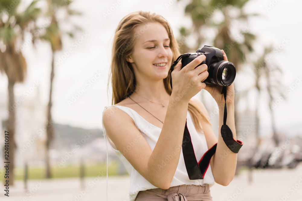 Pretty young girl laughing while taking pictures on professional dslr camera walking outdoors in the park on the summer sunny day, traveling, making photos, photography as a hobby, looking into camera