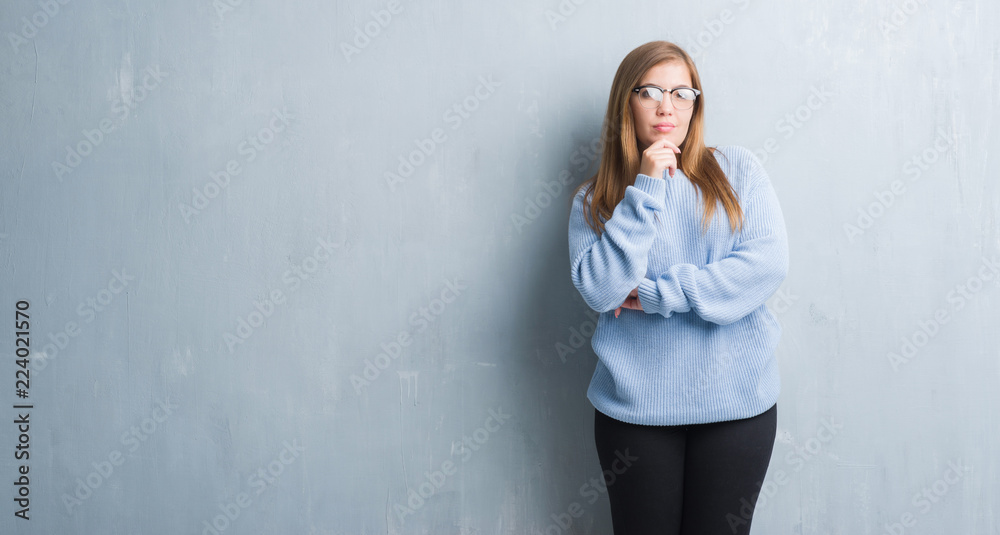 Young adult woman over grey grunge wall wearing glasses looking confident at the camera with smile with crossed arms and hand raised on chin. Thinking positive. Young adult woman over grey grunge wall wearing glasses looking confident at the camera with smile with crossed arms and hand raised on chin. Thinking positive.