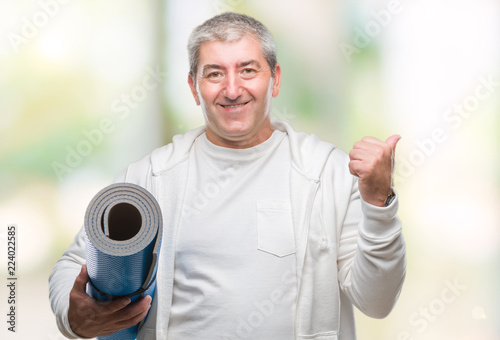 Handsome senior man holding yoga mat over isolated background pointing and showing with thumb up to the side with happy face smiling
