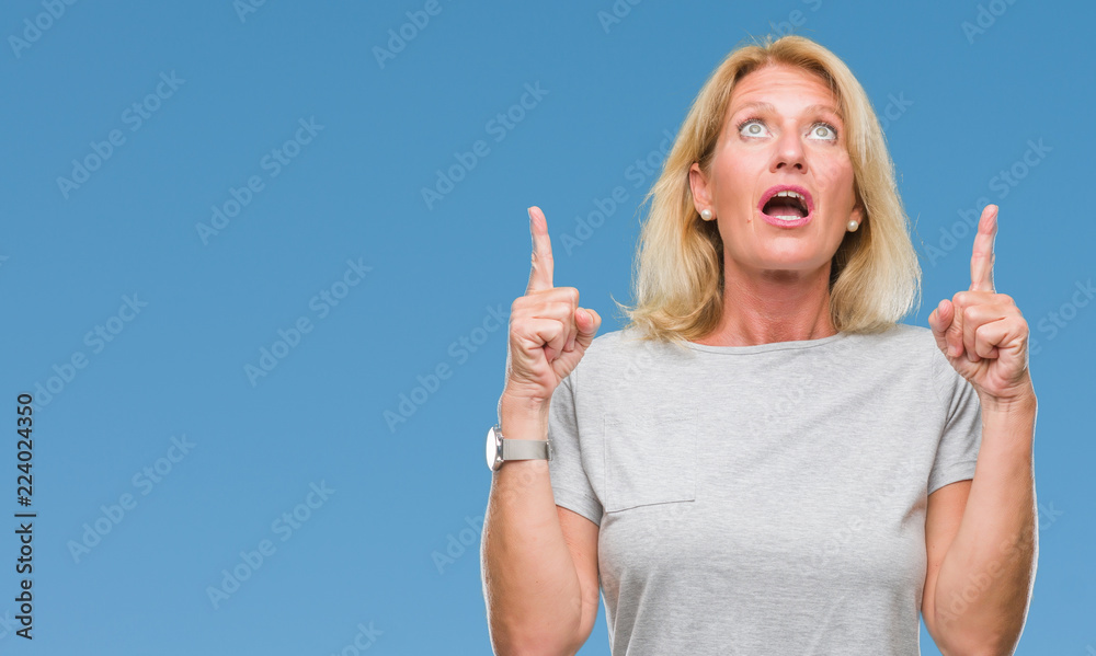 Middle age blonde woman over isolated background amazed and surprised looking up and pointing with fingers and raised arms. Middle age blonde woman over isolated background amazed and surprised looking up and pointing with fingers and raised arms.