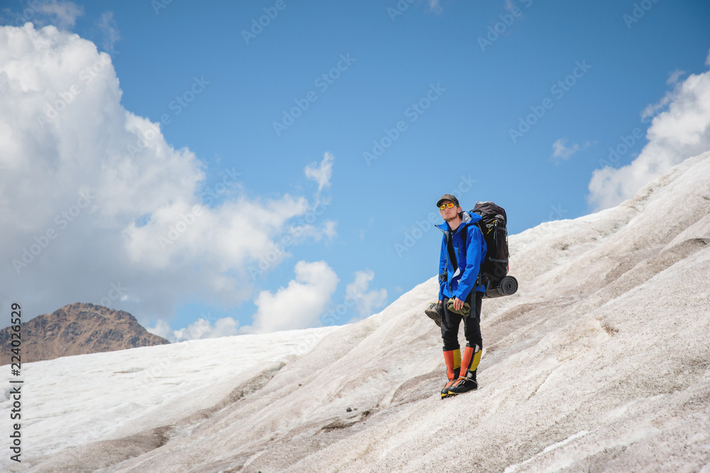 A mountaineer with a backpack walks in wheelchairs, stands on a dusty glacier with sneakers in the hands between cracks in the mountains