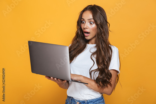 Portrait of shocked woman 20s with long hair surprising and holding gray laptop, isolated over yellow background