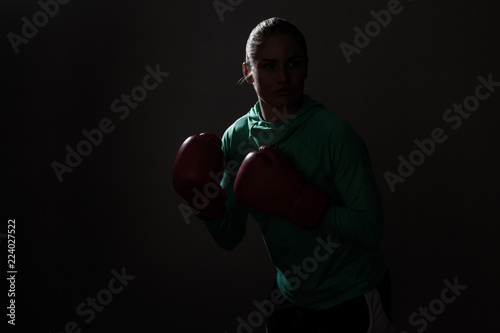 silhouette portrait of young boxer woman in green sport wear and red gloves standing in attack or defence position and looking away. indoor studio shot, isolated on black background. © khosrork