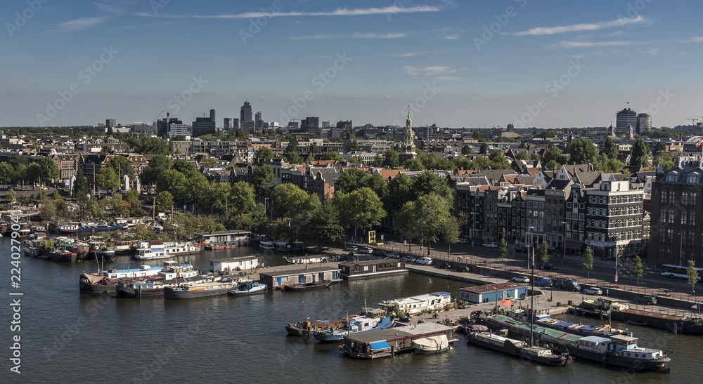 View north, over Amsterdam city, with many boats alongside the large canal. It is summertime and the sky is blue