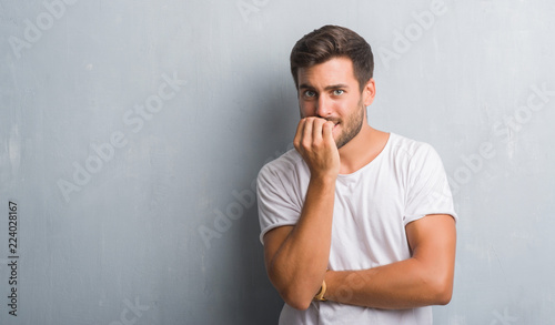 Handsome young man over grey grunge wall looking stressed and nervous with hands on mouth biting nails. Anxiety problem.