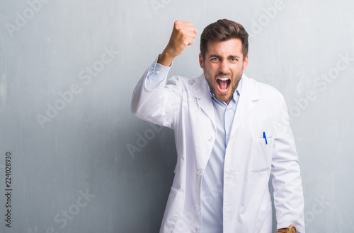Handsome young professional man over grey grunge wall wearing white coat angry and mad raising fist frustrated and furious while shouting with anger. Rage and aggressive concept. © Krakenimages.com