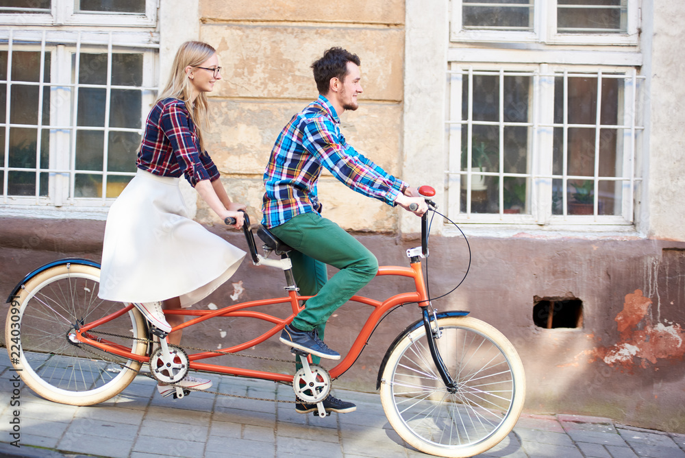 Young romantic pair, handsome man and pretty blond woman cycling together tandem double red bike along paved sidewalk on bright sunny summer day by old buildings Young romantic pair, handsome man and pretty blond woman cycling together tandem double red bike along paved sidewalk on bright sunny summer day by old buildings