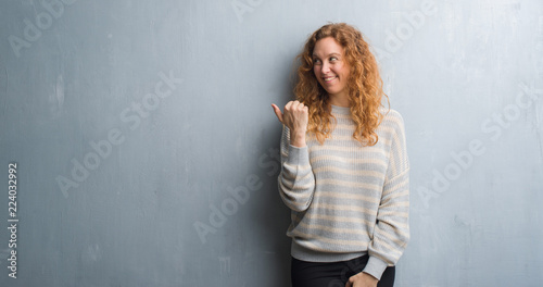 Young redhead woman over grey grunge wall smiling with happy face looking and pointing to the side with thumb up.