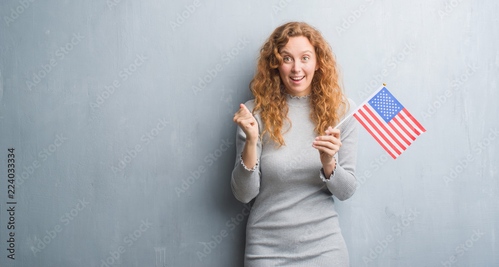 Young redhead woman over grey grunge wall holding flag of United States of America screaming proud and celebrating victory and success very excited, cheering emotion Young redhead woman over grey grunge wall holding flag of United States of America screaming proud and celebrating victory and success very excited, cheering emotion