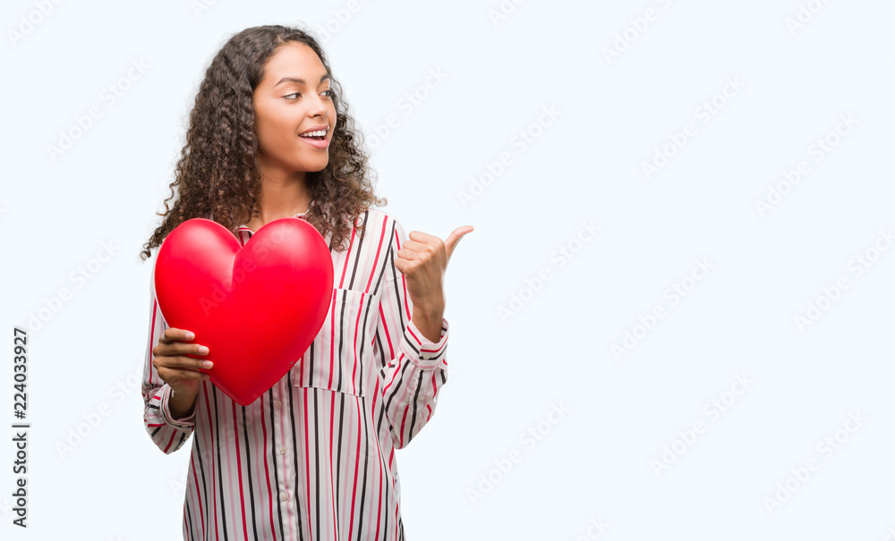 Young hispanic woman in love holding red heart pointing and showing with thumb up to the side with happy face smiling Young hispanic woman in love holding red heart pointing and showing with thumb up to the side with happy face smiling