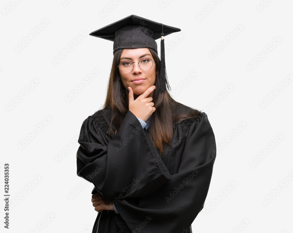 Young hispanic woman wearing graduated cap and uniform looking confident at the camera with smile with crossed arms and hand raised on chin. Thinking positive. Young hispanic woman wearing graduated cap and uniform looking confident at the camera with smile with crossed arms and hand raised on chin. Thinking positive.