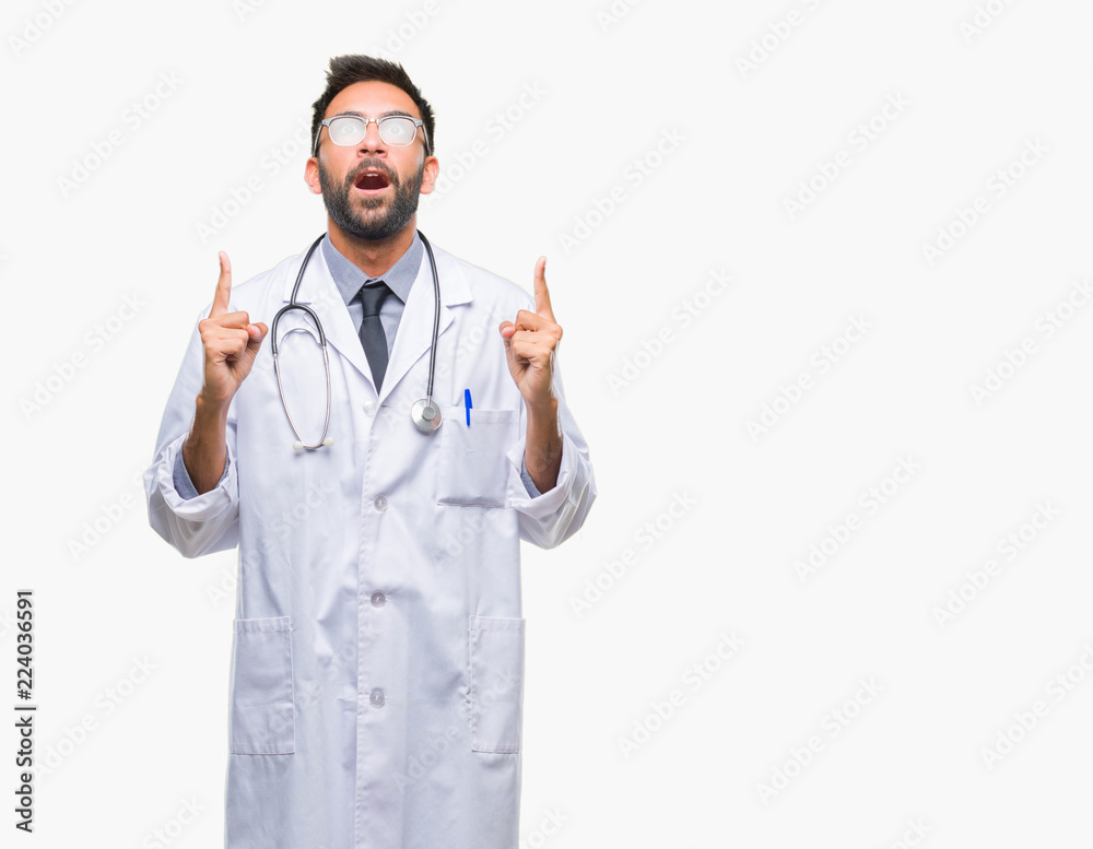 Adult hispanic doctor man over isolated background amazed and surprised looking up and pointing with fingers and raised arms. Adult hispanic doctor man over isolated background amazed and surprised looking up and pointing with fingers and raised arms.