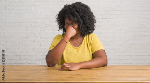 Young african american woman sitting on the table at home smelling something stinky and disgusting, intolerable smell, holding breath with fingers on nose. Bad smells concept.