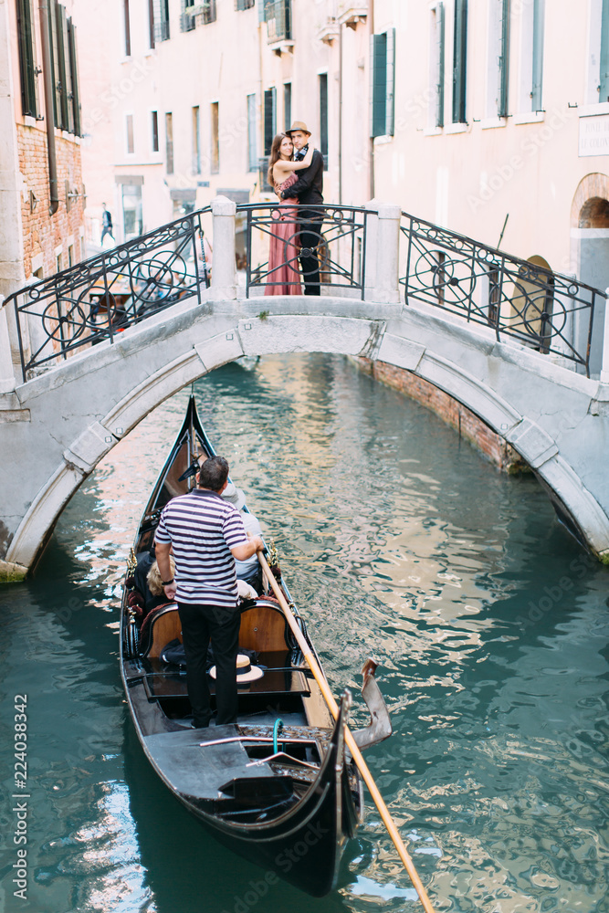 Venetian scenery with a gondola on a narrow canal. Romantic couple standing on the bridge of venetian canal.