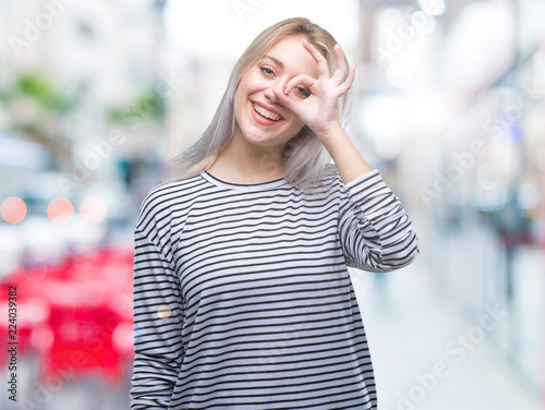 Young blonde woman over isolated background doing ok gesture with hand smiling, eye looking through fingers with happy face.