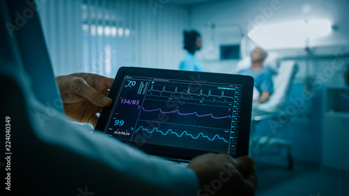 In the Hospital Doctor Holds Tablet Computer with Heart Rate and Other Health Data, In the Background Patients Lies in Bed while Nurse Checks Him.