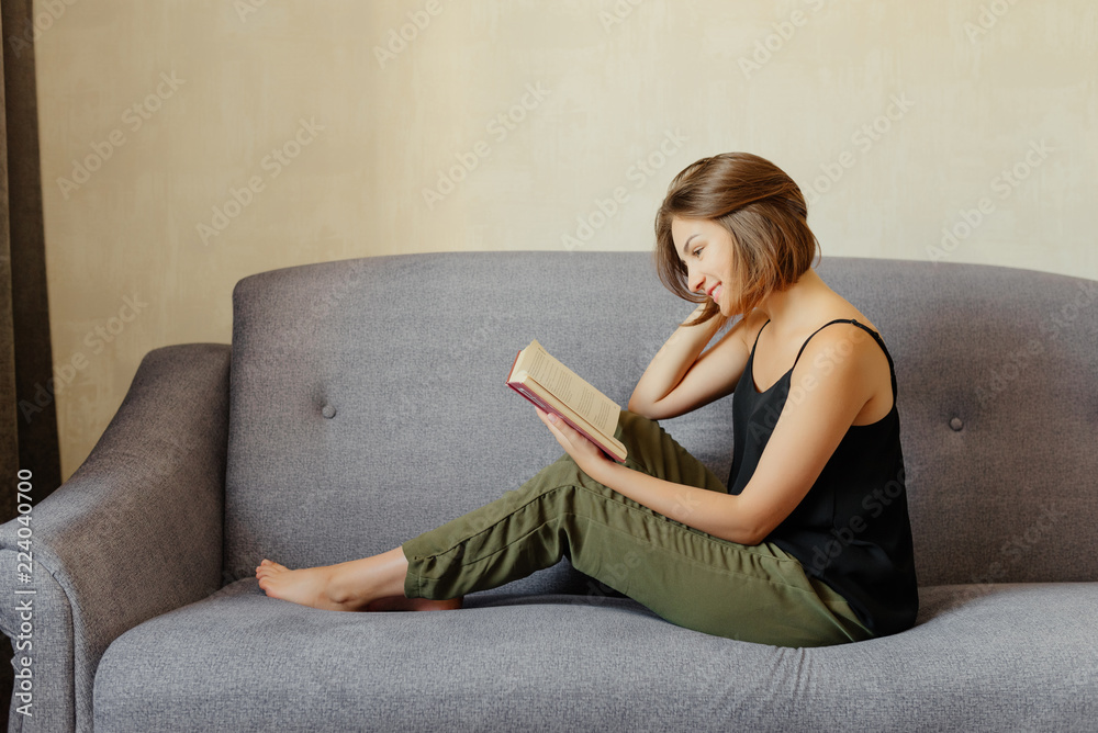 Side view of beautiful woman with short hair, sitting on the grey sofa and reading the book, at home.