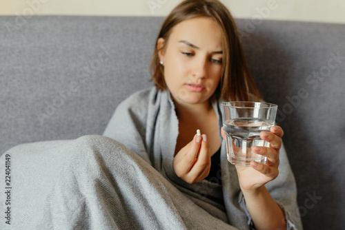 Woman have a cold, taking pills and holds a glass of water, sitting on the sofa, wrapped in the woolen blanket