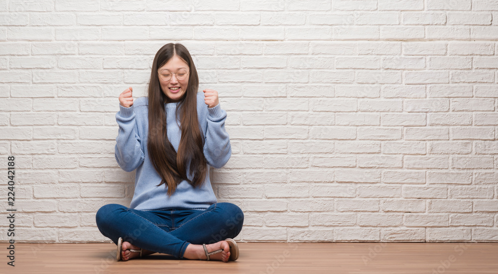 Young Chinese woman sitting on the floor over brick wall excited for success with arms raised celebrating victory smiling. Winner concept. Young Chinese woman sitting on the floor over brick wall excited for success with arms raised celebrating victory smiling. Winner concept.