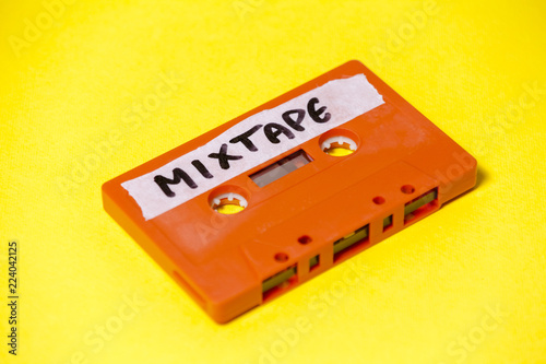 A vintage cassette tape (obsolete music technology), orange on a yellow surface, angled shot, carrying a label with the handwritten text Mixtape.
