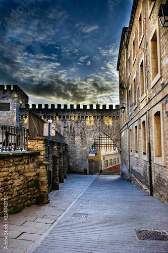 High dynamic range photograph of a street in the old part of Vitoria, Spain, with part of a crenellated wall and its raked door, cobblestone floor and granite stone facades