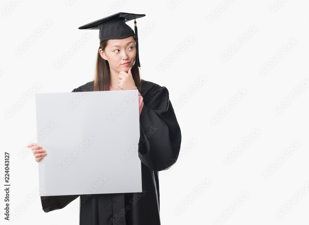 Young Chinese woman wearing graduate uniform holding banner serious face thinking about question, very confused idea Young Chinese woman wearing graduate uniform holding banner serious face thinking about question, very confused idea