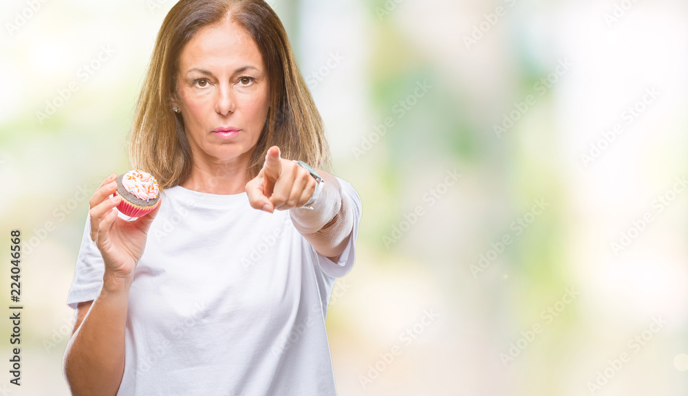 Middle age hispanic woman eating cupcake over isolated background pointing with finger to the camera and to you, hand sign, positive and confident gesture from the front Middle age hispanic woman eating cupcake over isolated background pointing with finger to the camera and to you, hand sign, positive and confident gesture from the front