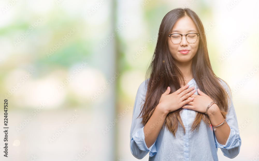 Young asian business woman wearing glasses over isolated background smiling with hands on chest with closed eyes and grateful gesture on face. Health concept.