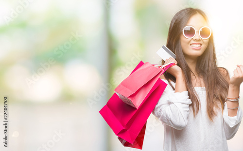 Young asian woman holding shopping bags on sales over isolated background screaming proud and celebrating victory and success very excited, cheering emotion
