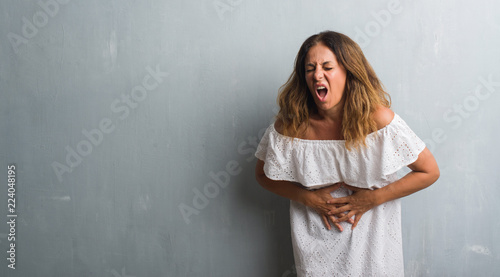 Middle age hispanic woman standing over grey grunge wall with hand on stomach because indigestion, painful illness feeling unwell. Ache concept.