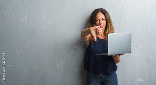 Middle age hispanic woman standing over grey grunge wall using laptop with angry face, negative sign showing dislike with thumbs down, rejection concept