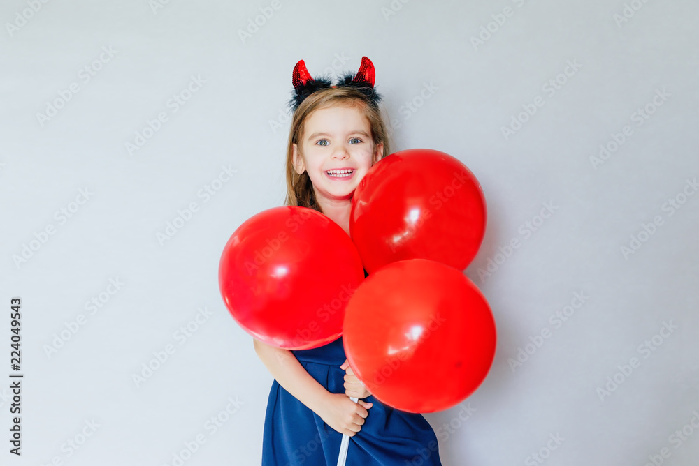 Happy funny girl with red balloons and red horns on her head near the empty gray concrete walls. Concept of the celebration. Halloween. Merry Christmas. Sale. Black Friday. Happy funny girl with red balloons and red horns on her head near the empty gray concrete walls. Concept of the celebration. Halloween. Merry Christmas. Sale. Black Friday.