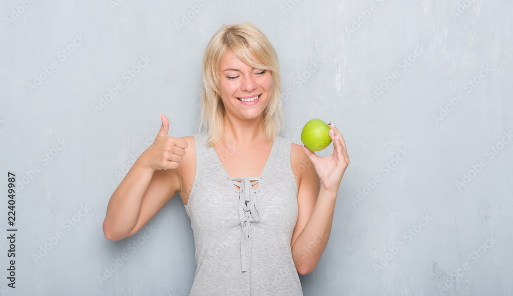 Adult caucasian woman over grunge grey wall eating green apple happy with big smile doing ok sign, thumb up with fingers, excellent sign Adult caucasian woman over grunge grey wall eating green apple happy with big smile doing ok sign, thumb up with fingers, excellent sign
