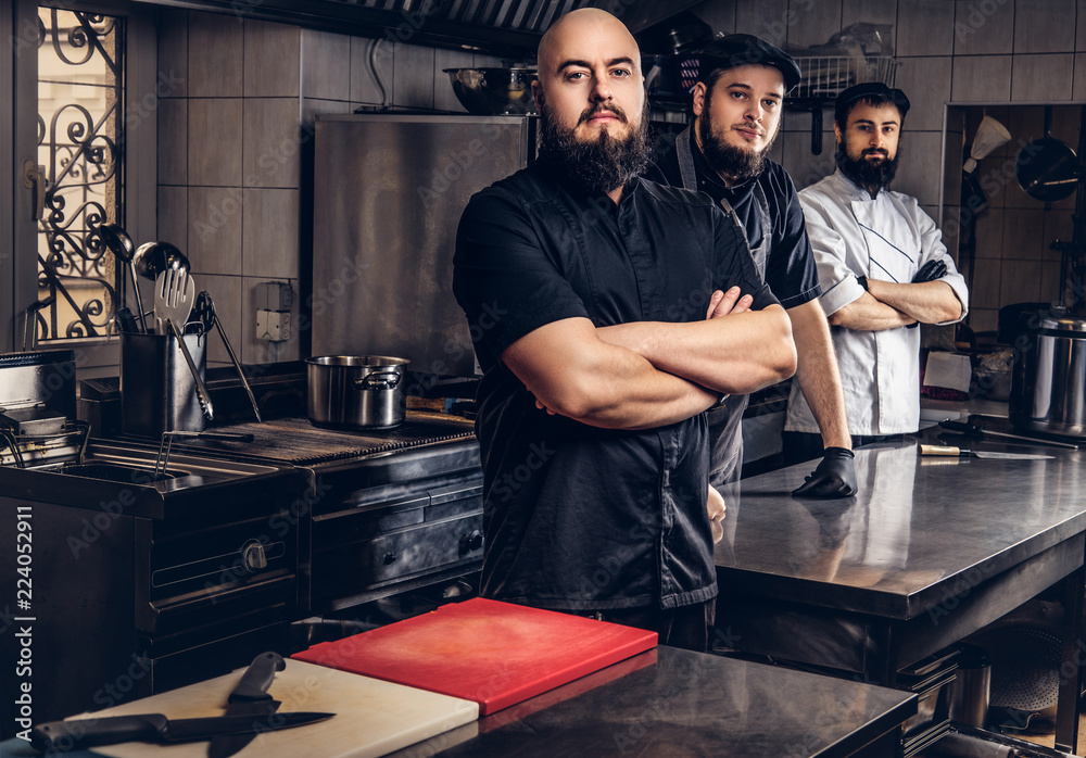 Team of professional bearded cooks dressed in uniforms standing in kitchen.