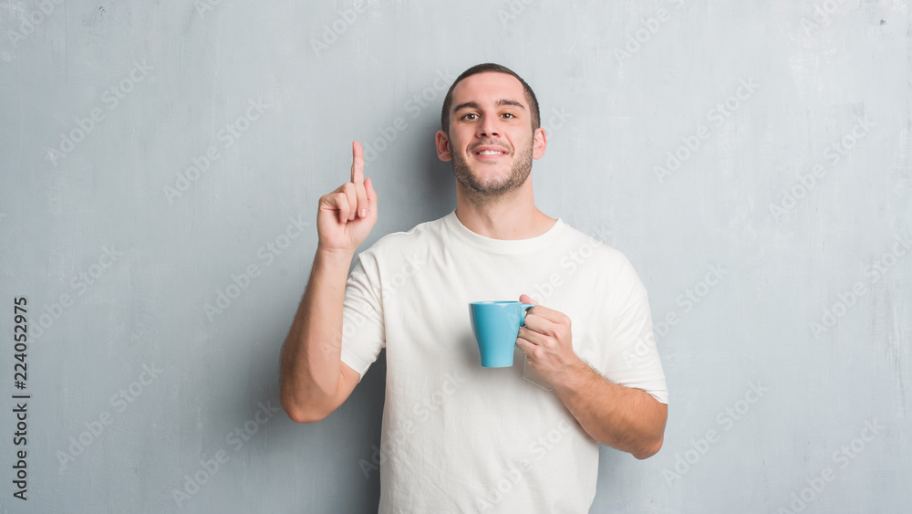 Young caucasian man over grey grunge wall driking a cup of coffee surprised with an idea or question pointing finger with happy face, number one Young caucasian man over grey grunge wall driking a cup of coffee surprised with an idea or question pointing finger with happy face, number one