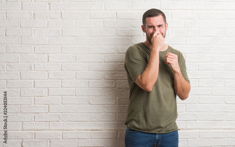 Young caucasian man standing over white brick wall smelling something stinky and disgusting, intolerable smell, holding breath with fingers on nose. Bad smells concept. Young caucasian man standing over white brick wall smelling something stinky and disgusting, intolerable smell, holding breath with fingers on nose. Bad smells concept.
