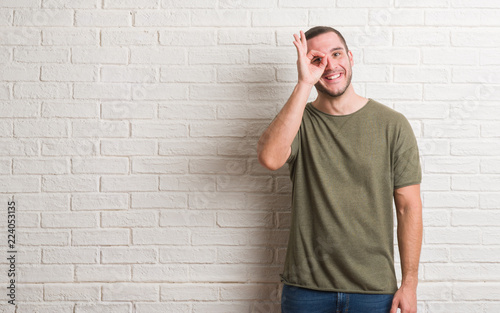 Young caucasian man standing over white brick wall doing ok gesture with hand smiling, eye looking through fingers with happy face.