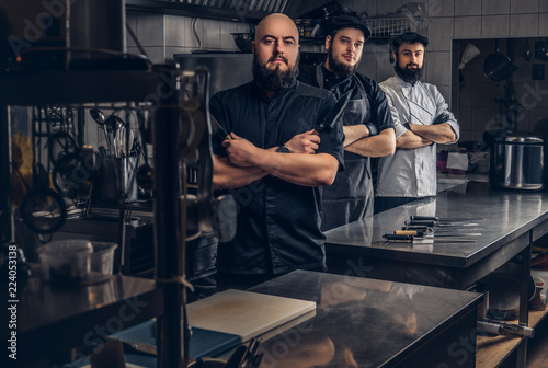 Team of professional bearded cooks dressed in uniforms posing with knives in kitchen.