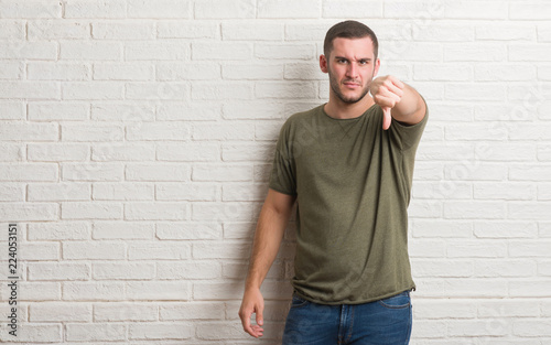 Young caucasian man standing over white brick wall looking unhappy and angry showing rejection and negative with thumbs down gesture. Bad expression.