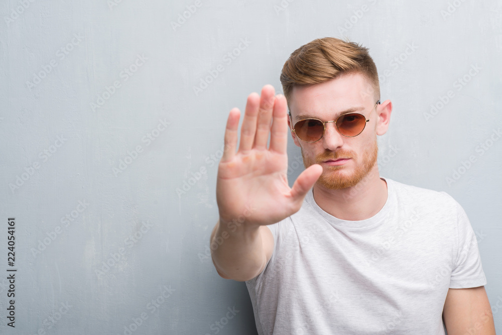 Young redhead man over grey grunge wall wearing retro sunglasses with open hand doing stop sign with serious and confident expression, defense gesture Young redhead man over grey grunge wall wearing retro sunglasses with open hand doing stop sign with serious and confident expression, defense gesture