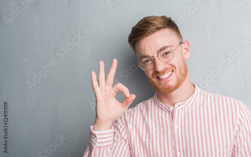 Young redhead man over grey grunge wall doing ok sign with fingers, excellent symbol