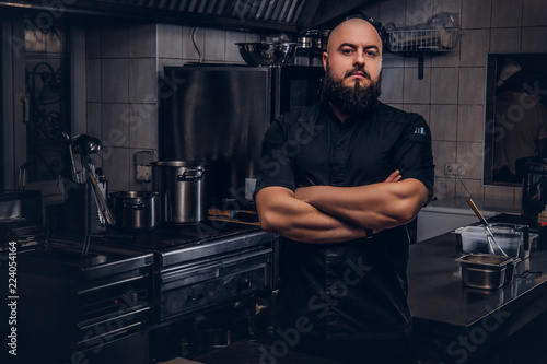 Brutal bearded chef cook in black uniform standing with crossed arms in the kitchen.