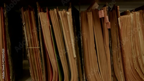 Camera movement along a file shelf. The paper of the files is old. In the background of the archive are even more shelves.