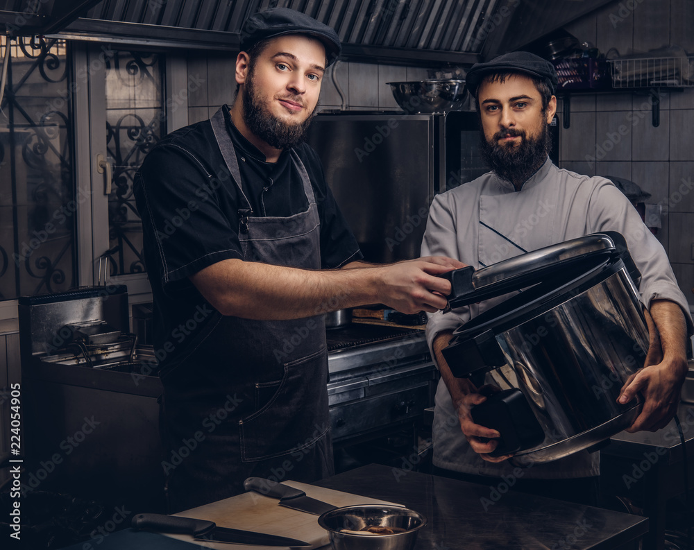 Two brutal cooks dressed in uniforms and hats standing with big multicooker in the kitchen. Two brutal cooks dressed in uniforms and hats standing with big multicooker in the kitchen.