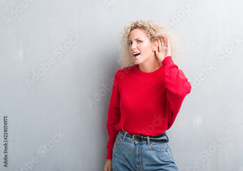 Young blonde woman with curly hair over grunge grey background smiling with hand over ear listening an hearing to rumor or gossip. Deafness concept.