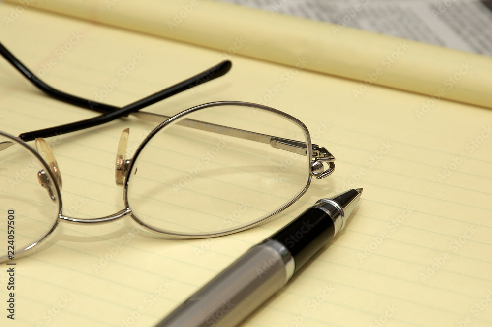 A yellow lined legal pad, reading glasses and a ballpoint pen in an office. A yellow lined legal pad, reading glasses and a ballpoint pen in an office.
