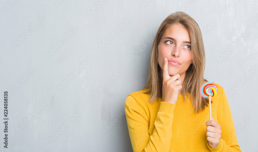 Beautiful young woman over grunge grey wall eating lollipop candy serious face thinking about question, very confused idea Beautiful young woman over grunge grey wall eating lollipop candy serious face thinking about question, very confused idea