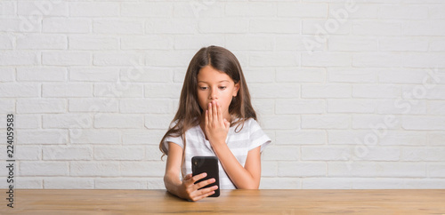 Young hispanic kid sitting on the table using smartphone cover mouth with hand shocked with shame for mistake, expression of fear, scared in silence, secret concept