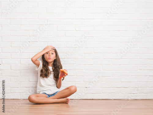 Young hispanic kid sitting on the floor over white brick wall eating pizza slice stressed with hand on head, shocked with shame and surprise face, angry and frustrated. Fear and upset for mistake.