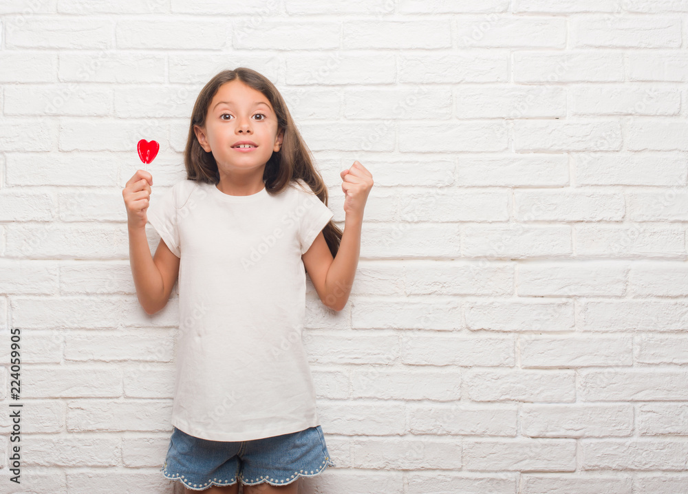 Young hispanic kid over white brick wall eating red heart lollipop candy screaming proud and celebrating victory and success very excited, cheering emotion Young hispanic kid over white brick wall eating red heart lollipop candy screaming proud and celebrating victory and success very excited, cheering emotion